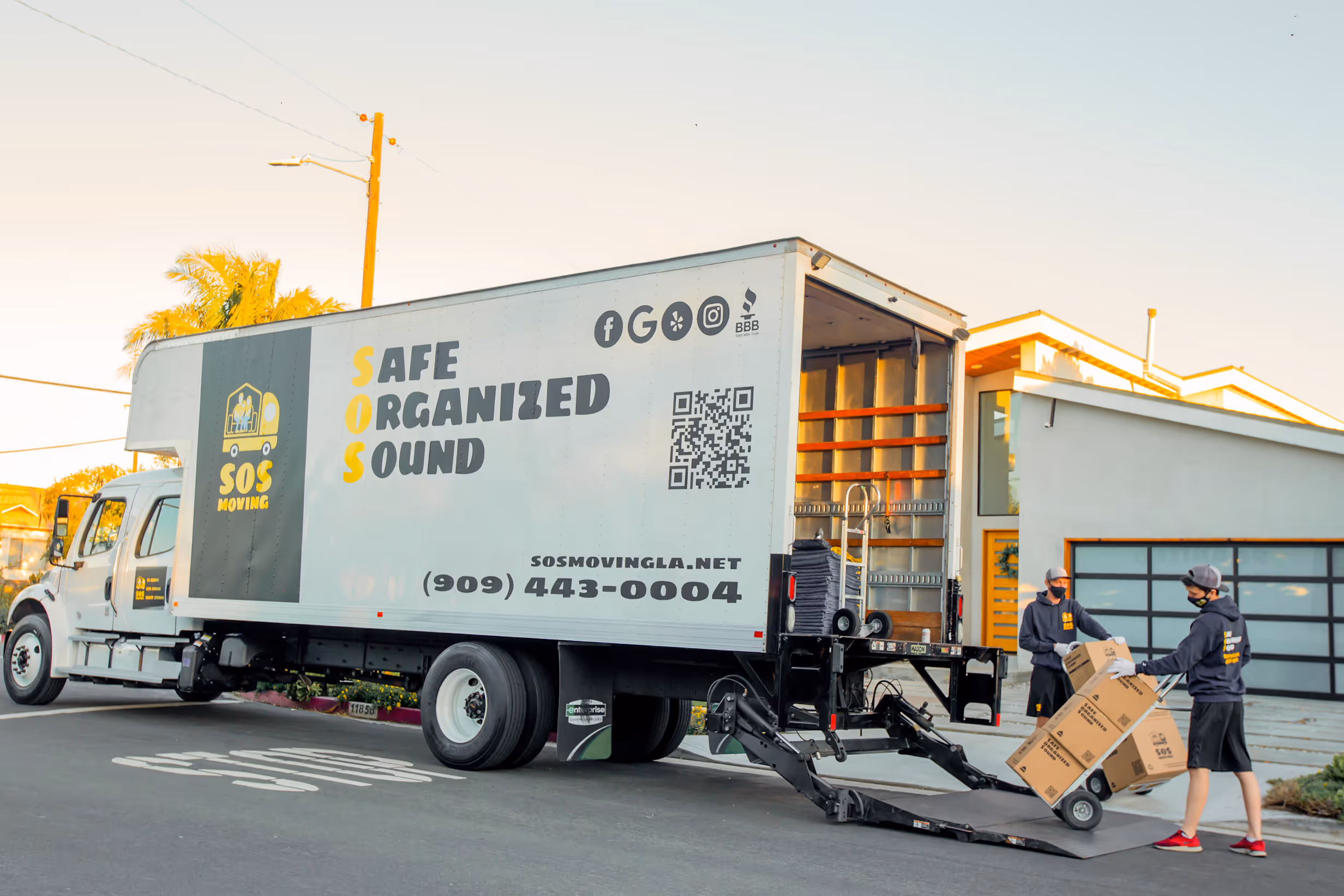 Two movers wearing masks unloading boxes on a hand truck from an SOS Moving truck outside a modern house.